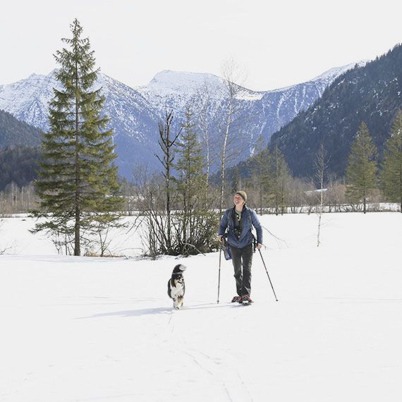 Eine Wanderin mit Schneeschuhen und Wanderstöcken in einer Schneelandschaft, von einem Hund begleitet.