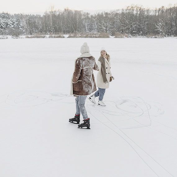 Zwei Frauen beim Schlittschuhlaufen auf einem zugeschneiten See, die sich dabei an den Händen halten
