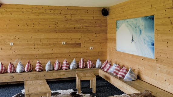 Gemütliche Lounge im alpenländlichen Stil, mit bunten Kissen, Holzwänden, Kuhfell auf dem Boden und kleinen Holzhockern.