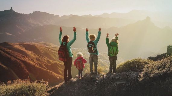 Eine Frau steht mit drei Kindern auf einem Berg. Alle haben die Hände nach oben hochgestreckt. Das Foto selbst zeigt eine Berglandschaft in Dämmerungsstimmung.