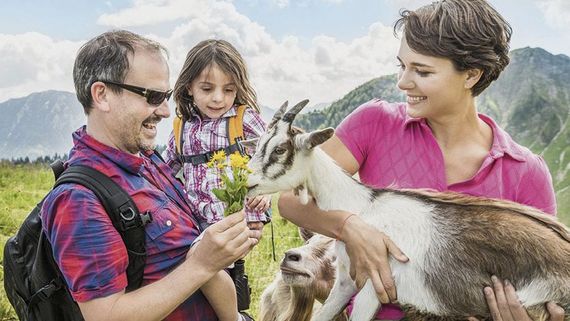Eine Familie in der freien Natur auf einer Bergwiese. Die Frau hat eine Ziege auf dem Arm, der Mann hält ihr Blumen zum Fressen hin.