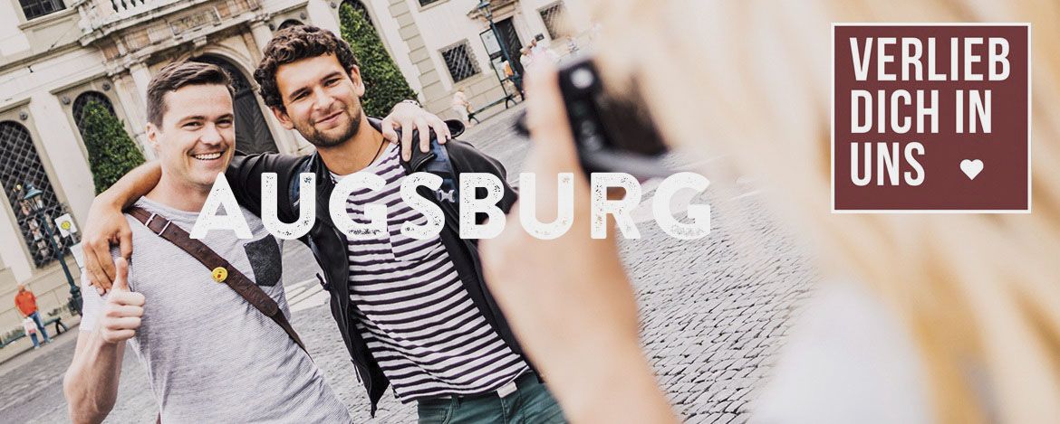 Zwei junge Männer lassen sich von einer Frau fotografieren. Im Hintergrund ist eine historische Hausfassade zu erkennen. Das Bild wird überlagert von den Schriftzügen "Verlieb dich in uns" und "Augsburg".