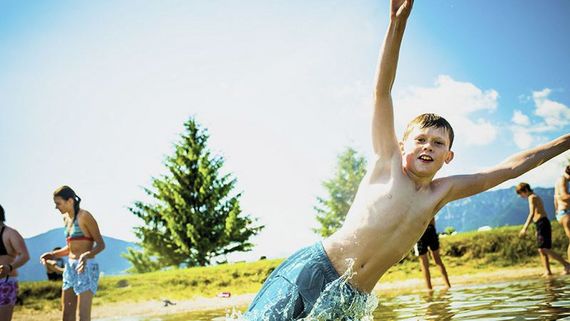 Badegäste in einem See, ein Kind im Zentrum des Bildes springt zur Seite ins Wasser