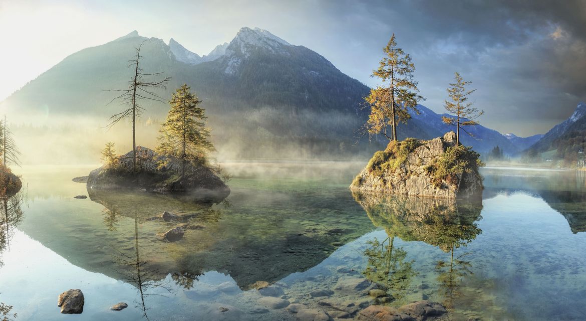 Ein künstlerisches Foto von einem See mit einem Felsmassiv im Hintergrund. Aus dem See ragen zwei kleinere Felsen mit einigen wenigen Nadelbäumen heraus. Über dem See ziehen dichte Nebelschwaden.